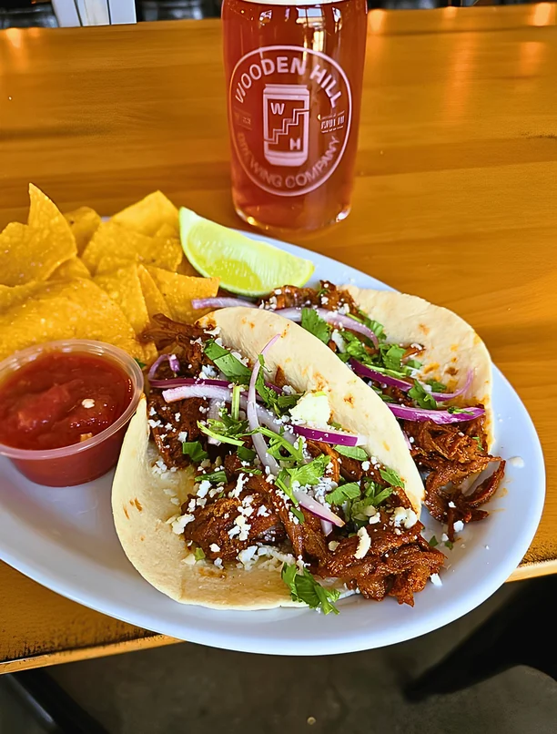 Two barbacoa tacos on a plate with a side of chips and salsa and a lime wedge. A beer is on the table in the background.