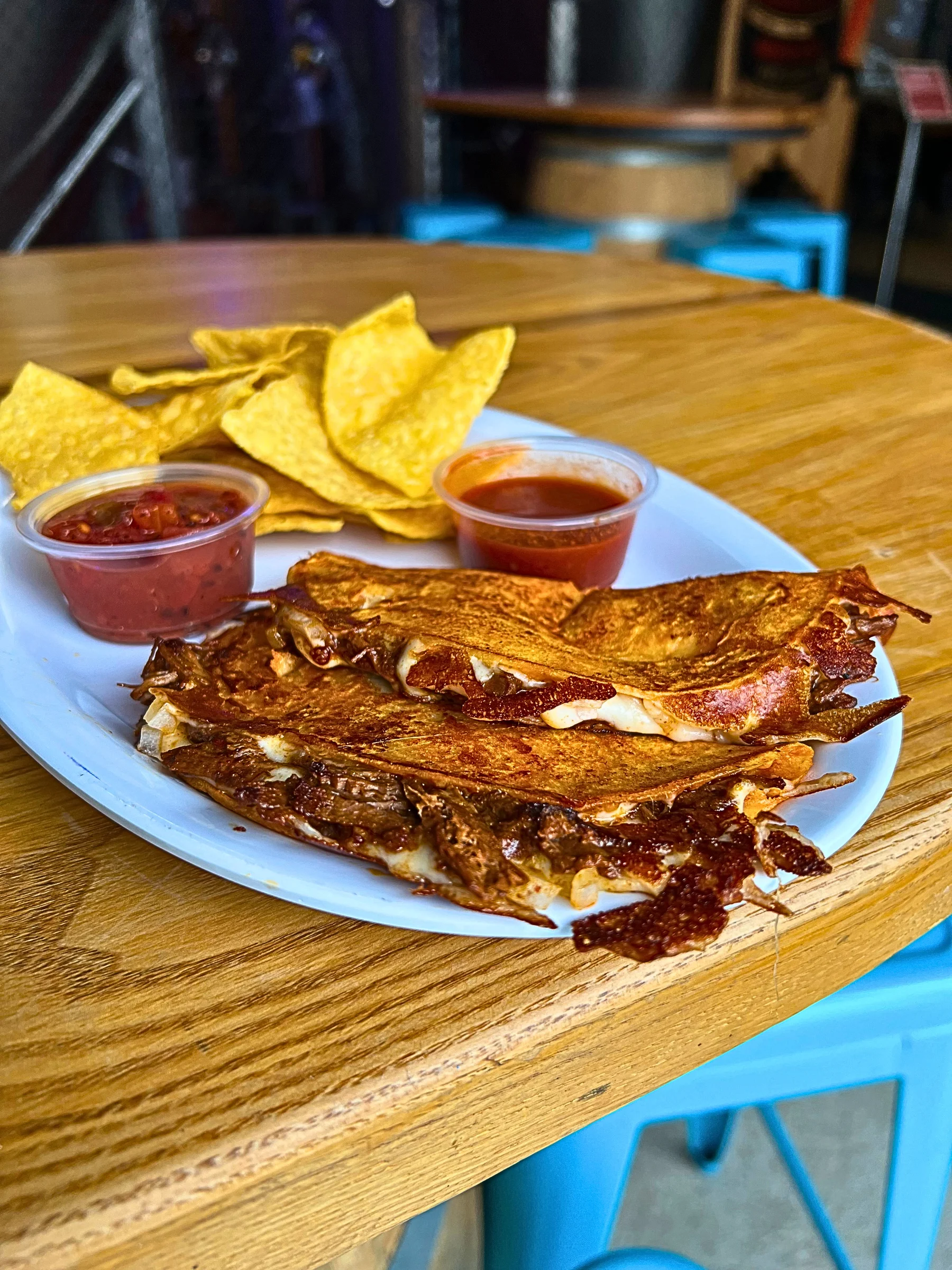 A close-up of a white plate on a wooden taproom table featuring two crispy Quesabirria Tacos. The consommé-dipped corn tortillas are pan-fried to a dark golden brown and overflowing with tender shredded birria beef, melted cheese, and diced onions. The tacos are served alongside yellow tortilla chips, a small cup of red salsa, and a side of rich consommé for dipping.