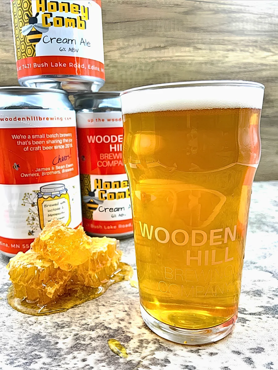 A glass of Honey Comb with a piece of honey comb and 3 stacked crowlers of the beer in the background.