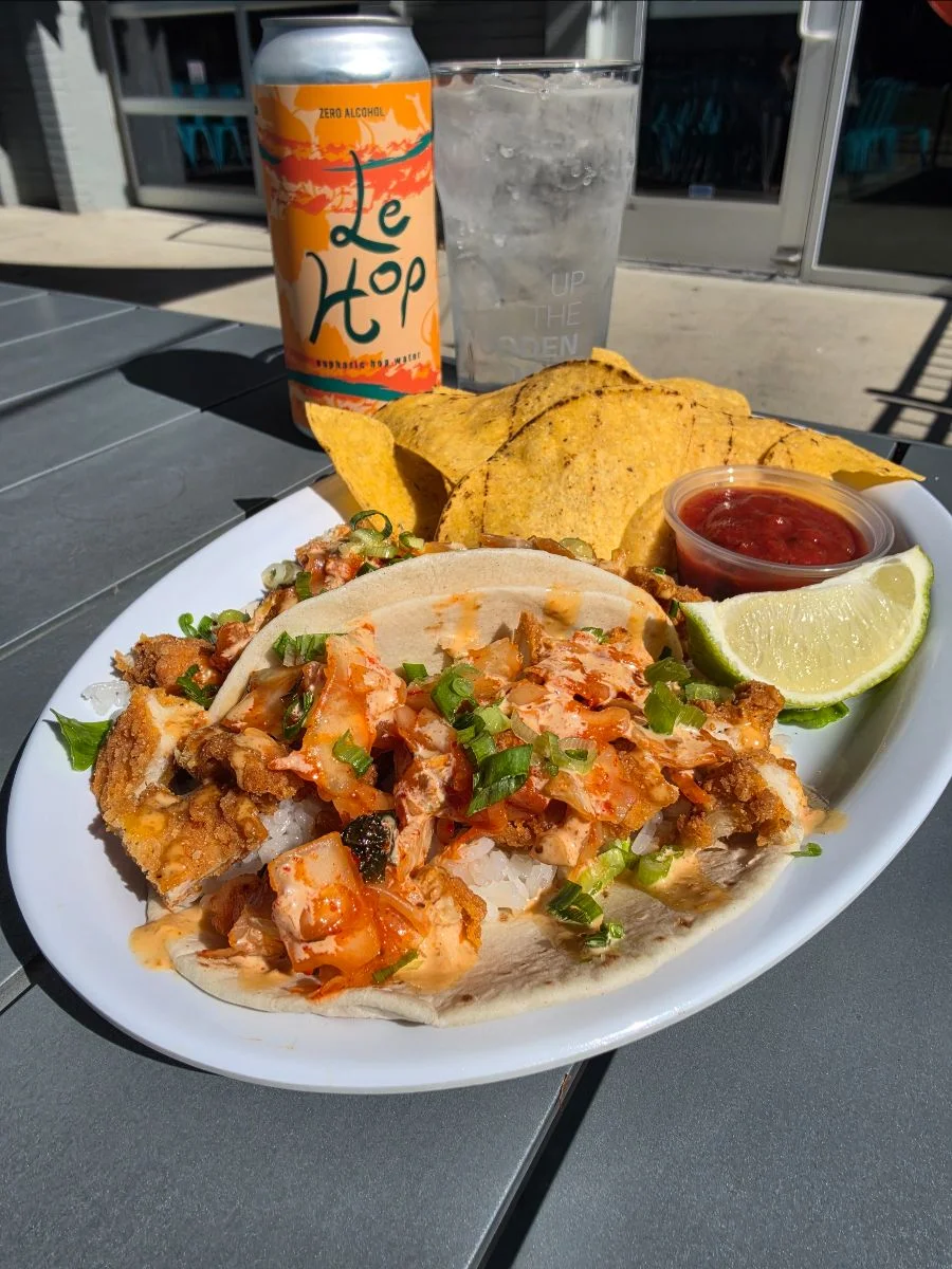 A plate with two Kimchi Kruncher Tacos with a side of chips and salse and a lime wedge. A can and glass of Le Hop in the background.