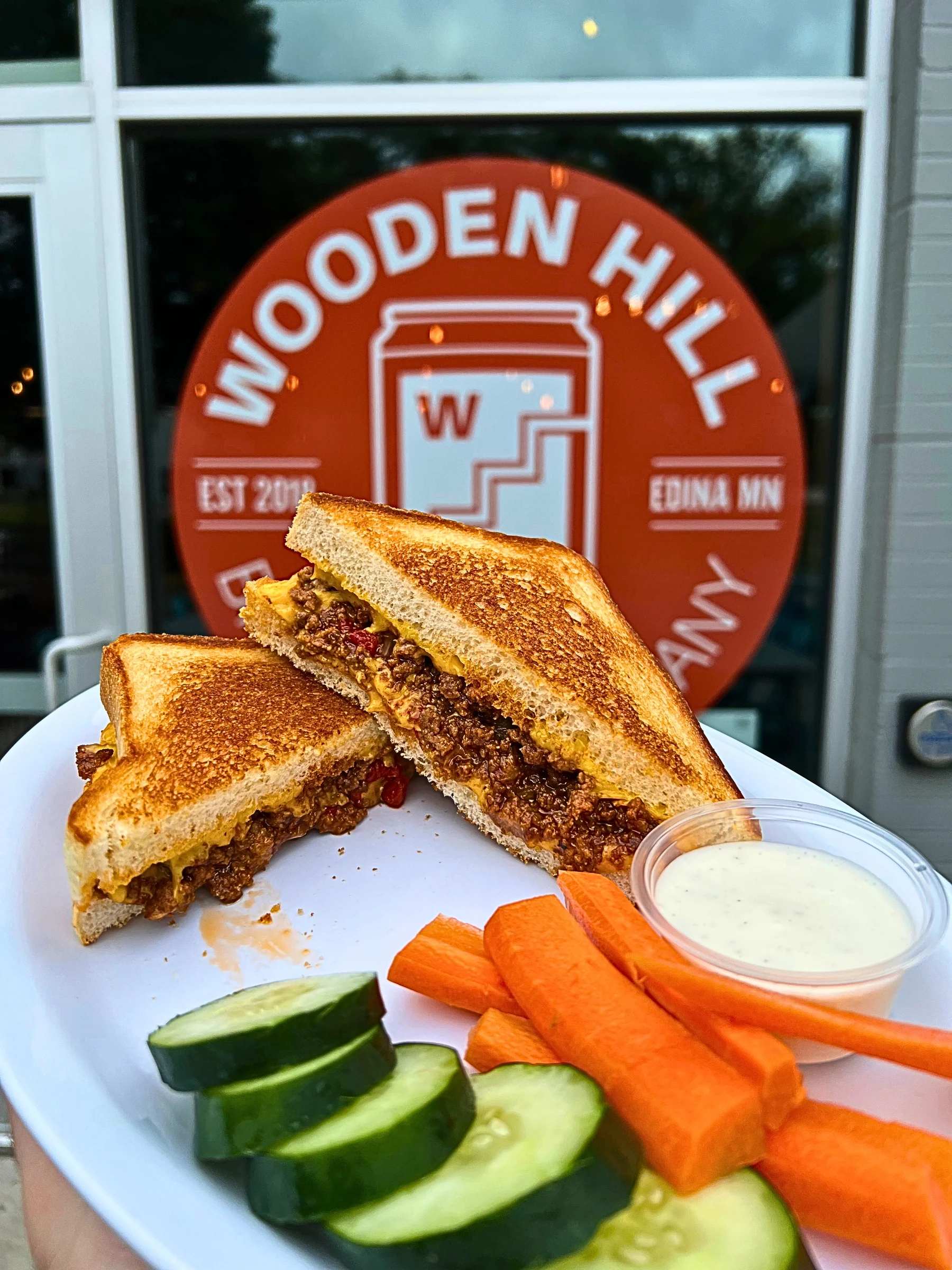 A white plate held up in front of a window featuring the orange Wooden Hill Brewing Company logo. On the plate is a Loaded Grilled Cheese sandwich cut in half, revealing thick, golden-brown Texas Toast stuffed with a generous portion of savory Sloppy Joe beef and melted cheddar cheese. It is served with a fresh side of cucumber slices, thick-cut carrot sticks, and a small cup of creamy dipping sauce.