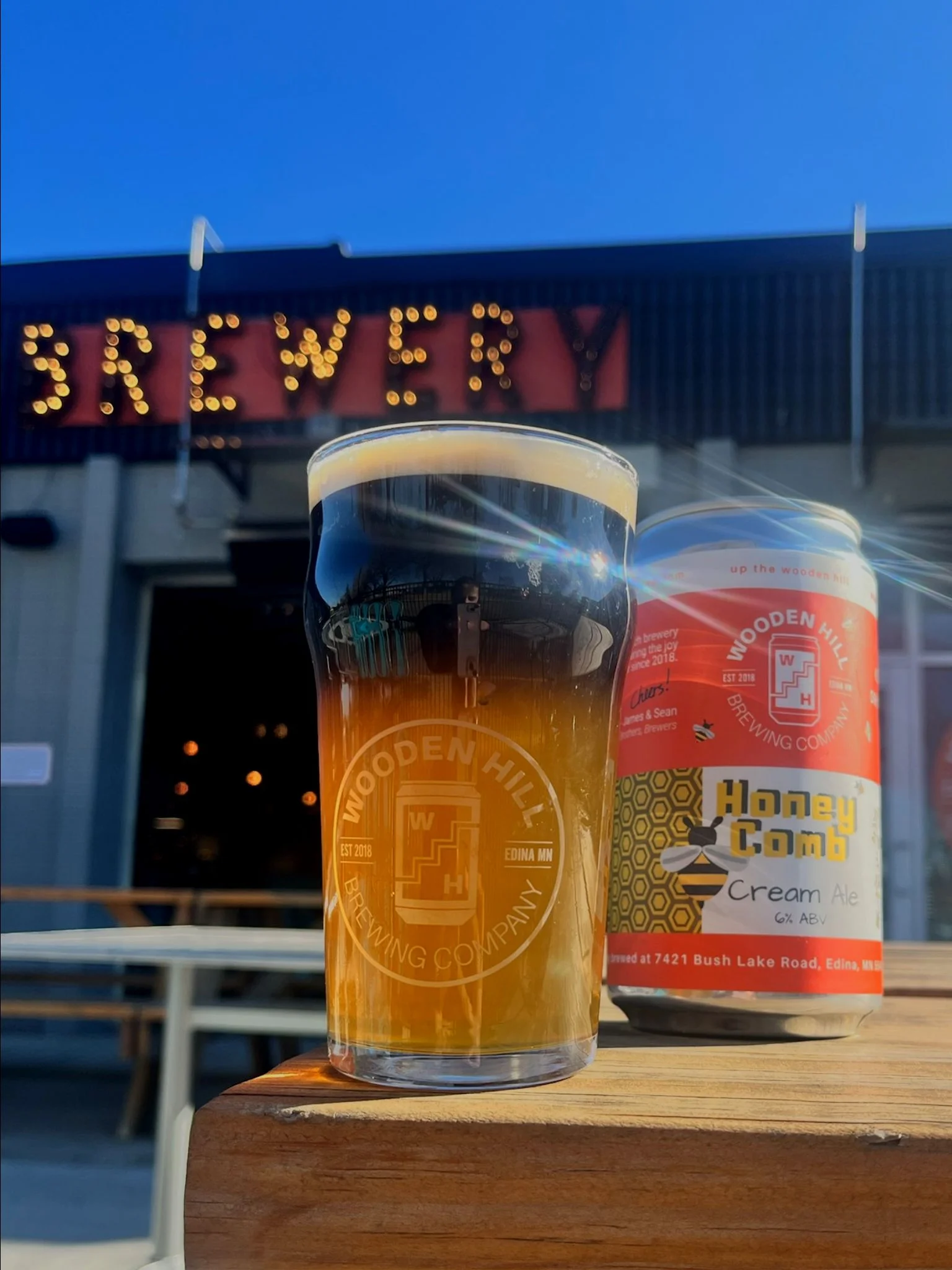 A glass of Honey Comb with a piece of honey comb and 3 stacked crowlers of the beer in the background.