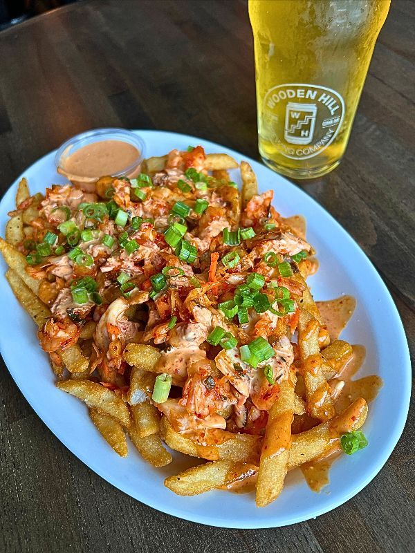 A close-up shot of Kimchi Fries served on a white oval plate resting on a dark wooden table. A generous mound of crispy seasoned fries is topped with vibrant garlic and red pepper kimchi, heavily drizzled with a creamy house soy harissa sauce, and garnished with freshly chopped green onions. A small side cup of extra sauce sits on the edge of the plate, and a cold pint of beer in a glass featuring the Wooden Hill Brewing Company logo is visible in the top right corner.