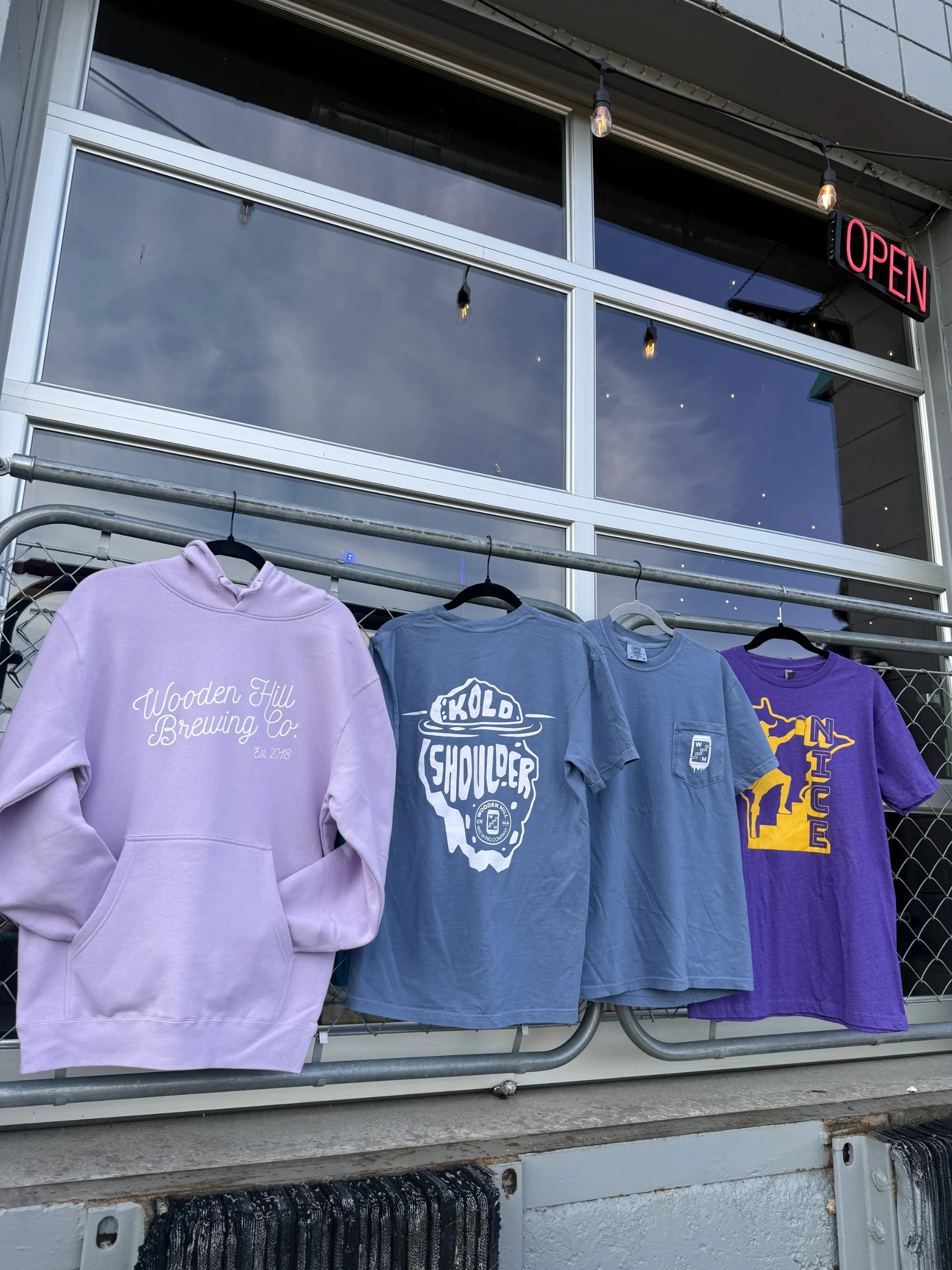 4 shirts hanging on a fence. Lavender Wooden Hill cursive sweatshirt, Kold Shoulder T-shirt (back), Kold Shoulder t-shirt (front), MN Nice tshirt. Fron left to right.