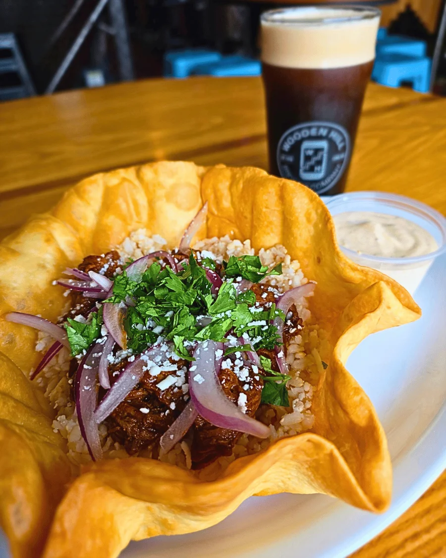 A fried flour tortilla bowl with rice, birria beef, red onions, queso fresco and cilantro on top, on a plate with a side of sour cream.