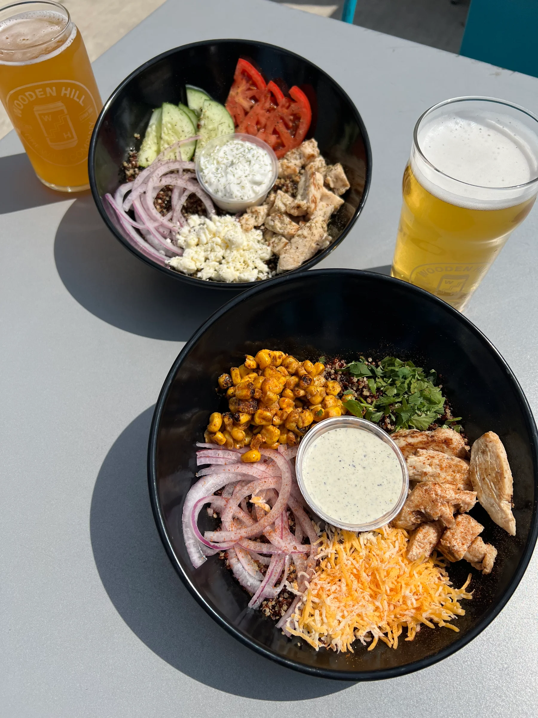 An overhead shot of two loaded grain bowls served in black dishes on a wooden table. The top bowl is the Athena Grain Bowl, featuring a base of multicolored garlic quinoa topped with sliced grilled chicken, crumbled feta cheese, fresh cucumbers, tomatoes, red onion, a dusting of Greek seasoning, and a small center cup of tzatziki. The bottom bowl is the Southwest Grain Bowl, loaded with multicolored garlic quinoa, grilled chicken, fire-roasted corn, shredded cheese, fresh cilantro, red onions, taco seasoning, and a center cup of creamy jalapeño ranch.