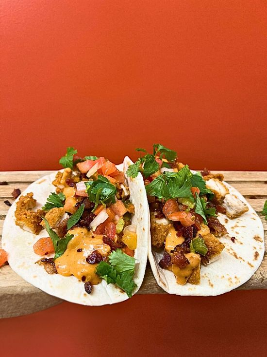 A close-up shot of two Crispy Queso Tacos served open on soft flour tortillas, resting on a rustic wooden surface against a solid, warm orange background. The tacos are loaded with chopped, golden-brown crispy chicken and heavily drizzled with a creamy, rich queso. They are finished with dark, crunchy bacon bits, fresh diced pico de gallo, and vibrant green cilantro leaves scattered over the top.