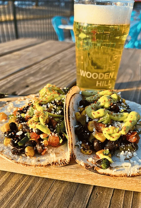 Two cowboy caviar tacos with Black Beans, Roasted Corn, Roasted Garlic, Tomato, Bell Pepper, Jalapeño & Cilantro |Crumbled Queso Fresco | Avocado Crema Chili Lime Seasoning and a beer in the background