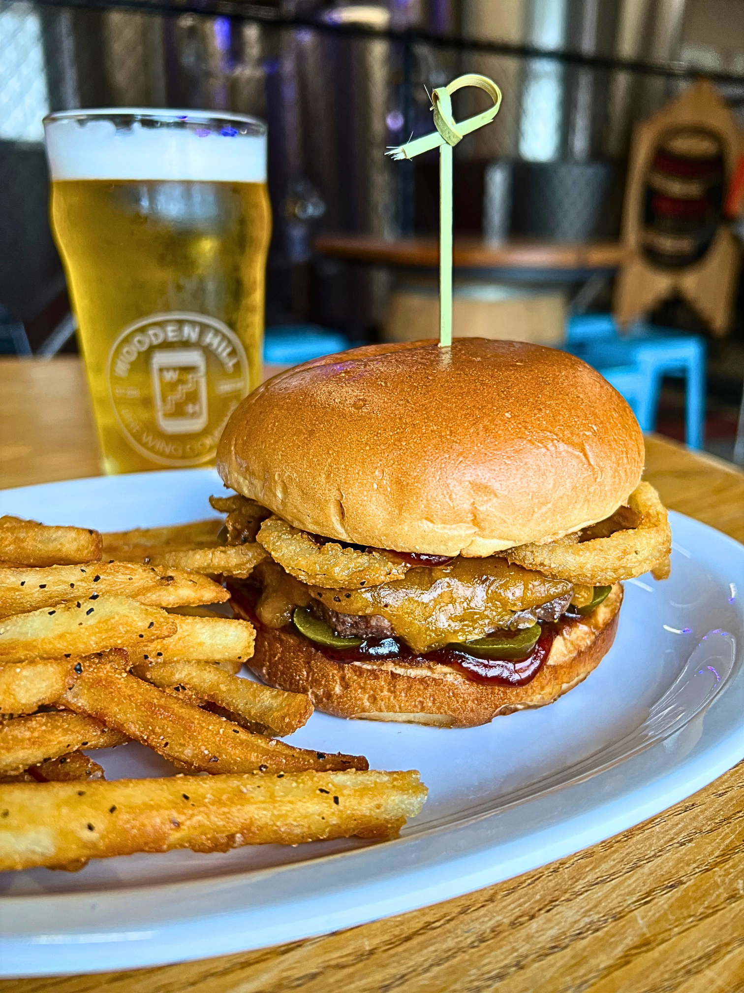 A cowboy burger on a white plate with a side of fried and a beer in the background.