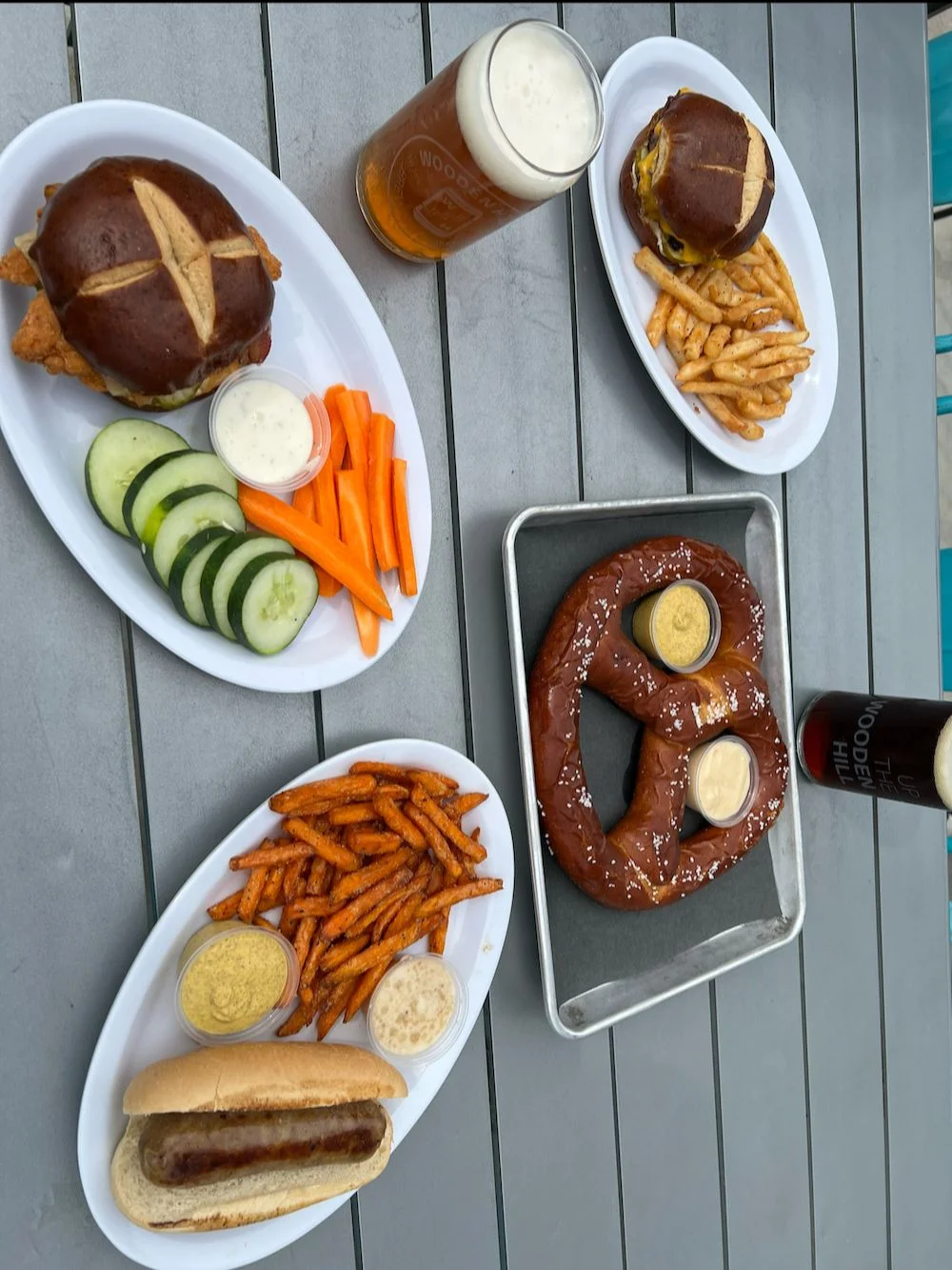 A table with multiple plates. two of a pretzel bun sandwich, one brat, and one bavarian pretzel, alongside two glasses of beer