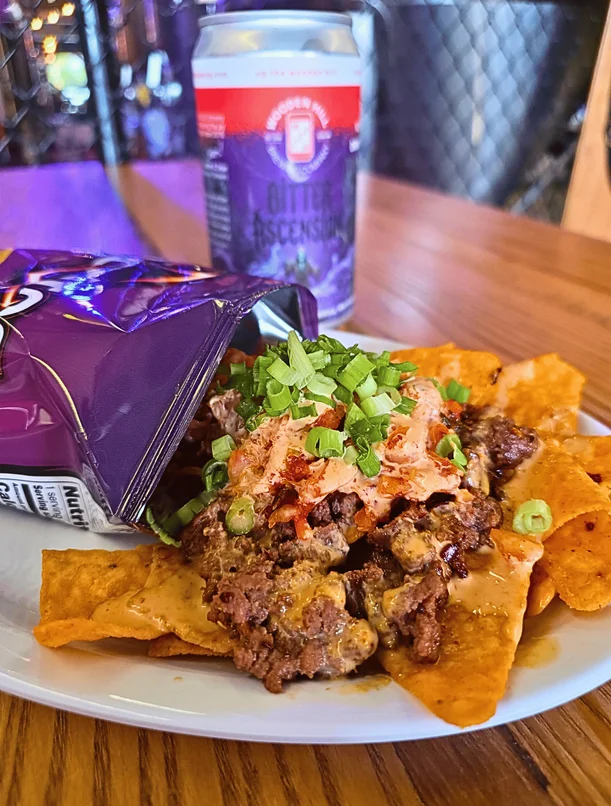 what looks like a walking taco spilled on a plate. A plate of ships with ground beef and toppings with a bag of doritos laying next to it and a crowler can in the background.