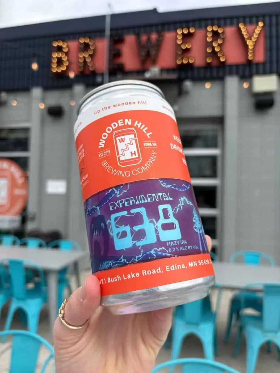 a photograph of a hand holding a big can of beer with an orange and purple label that reads "experimental 638" in front of a light up sign that says "brewery"