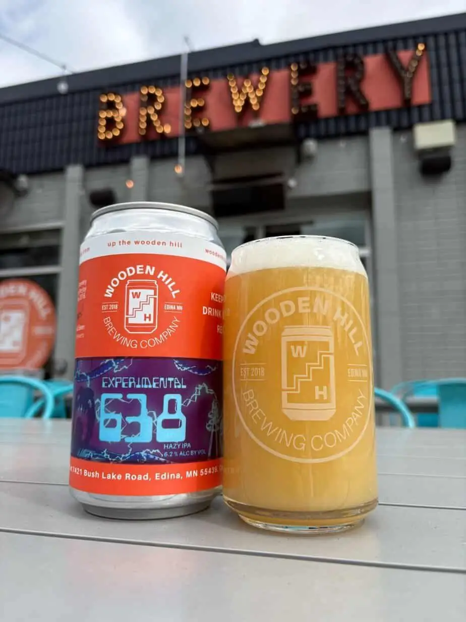 photograph of a big can of beer with an orange and purple label next to a pint of hazy beer, in front of a light up "Brewery" sign