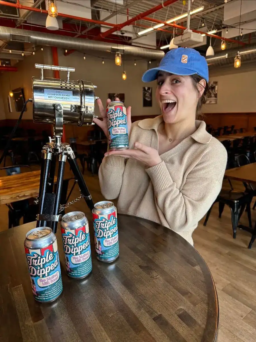 Picture of Mia holding a can of Triple Dipped showcasing her can artwork after winning the 2025 Aluminate Award for Best Can Design. The label is inspired by a retro old fashioned diner, with red and white banner stripes.