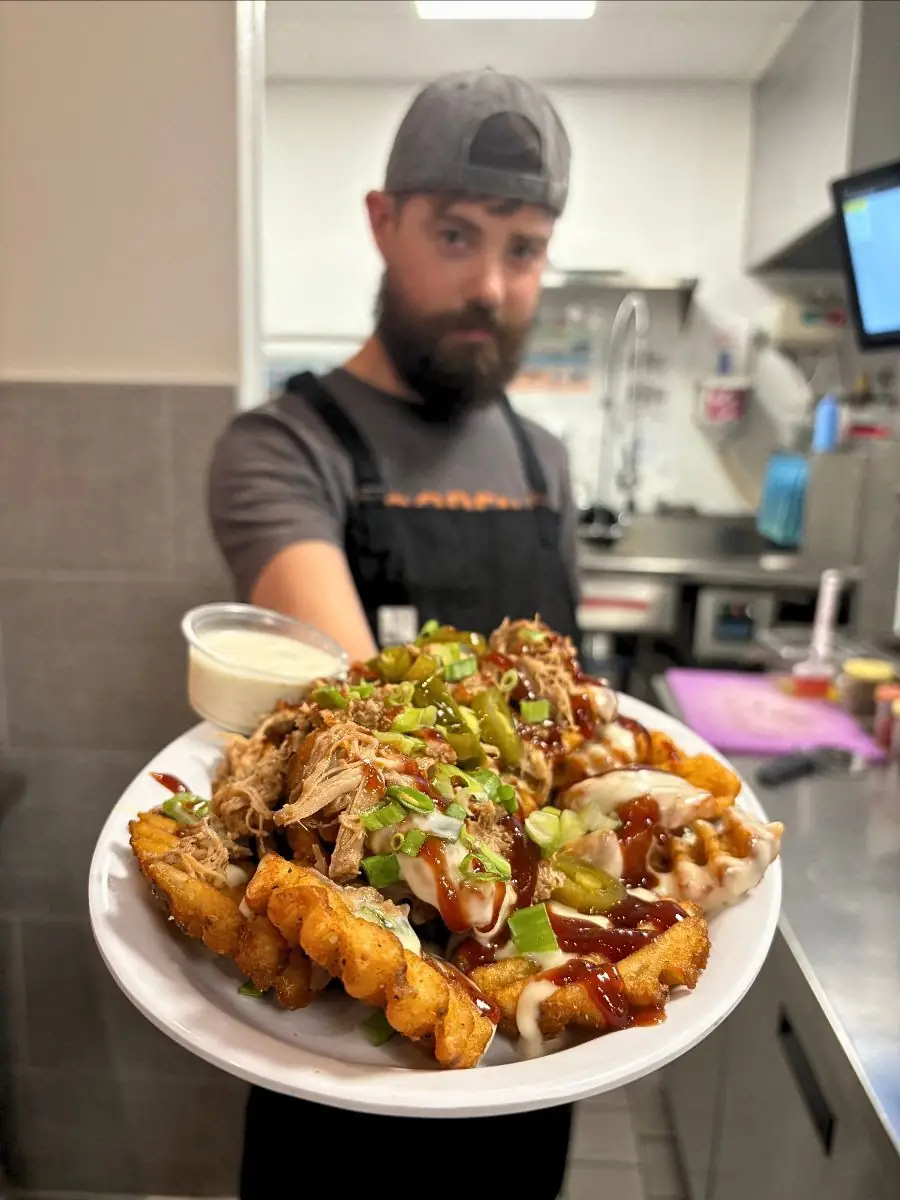 Image is focused on idaho nachos on a plate being held by a guy in the background in a kitchen.