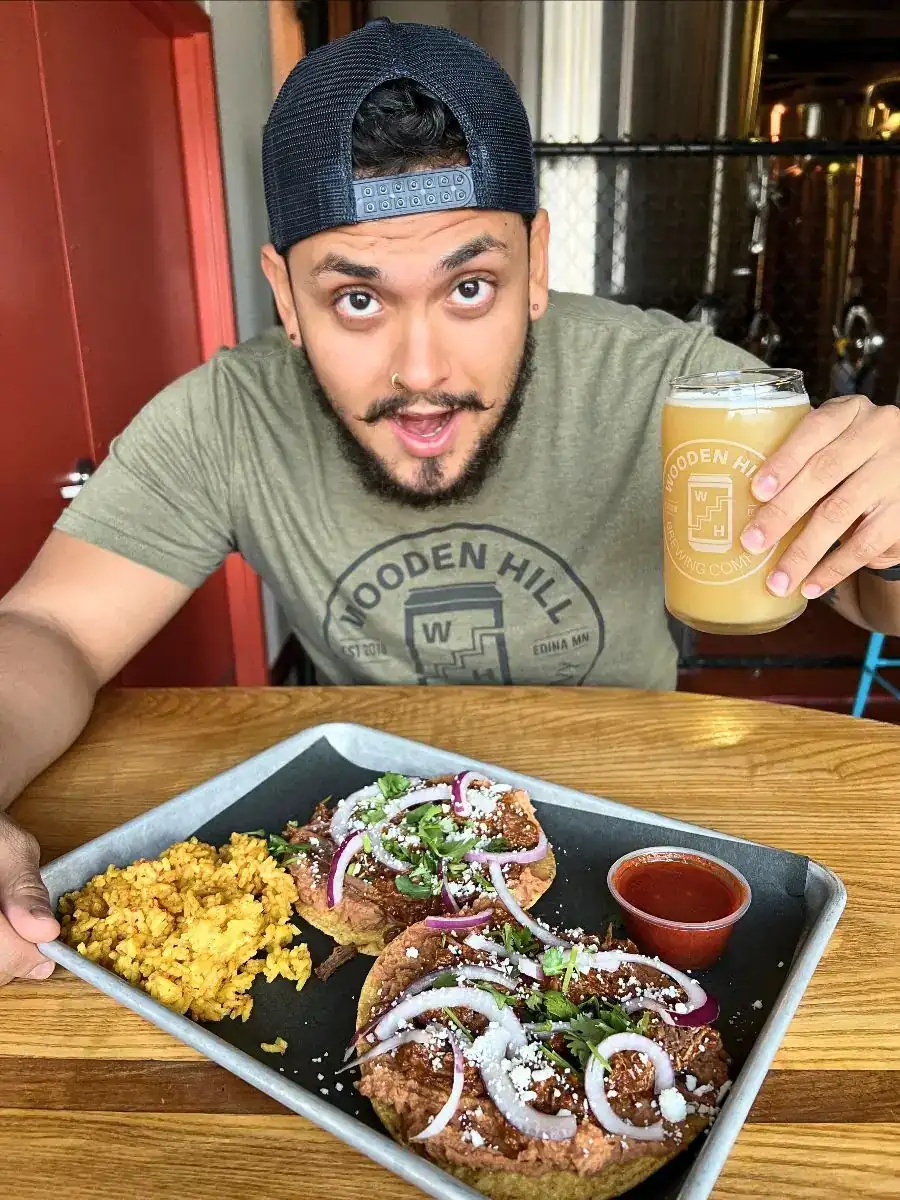 A guy holding a beer with an expressive face about the plate on the table in front of him. Plate has 2 crispy birria tostadas on it with spanish rice and a red consomme dipping sauce.