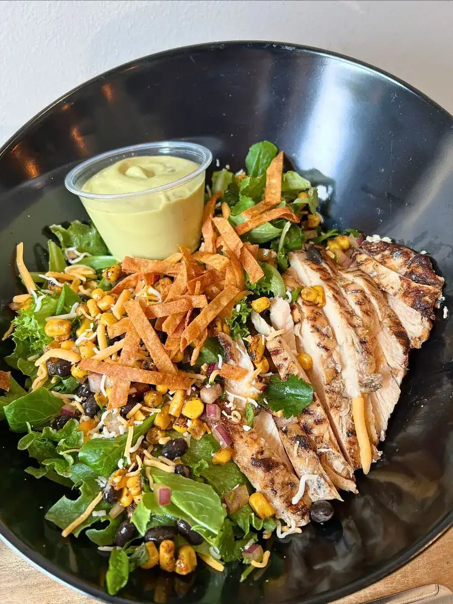 Southwest salad in a black bowl with a green dipping sauce in a clear container. Mixed green, cowboy caviar, shredded cheese, crispy tortilla strips, and grilled chicken.