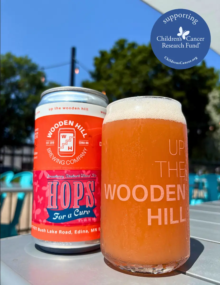 Image of a lighter beer in a wooden hill branded glass with the Hops for a Cure can (pink and blue logo with butterflies) behind it. Supporting Childrens Cancer Research Fund sticker at the top of the image. 
