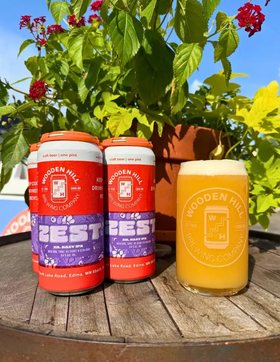 ZEST XII 4 pack (Wooden Hill orange cans with ZEST XII logo in shades of purple) of cans and a Wooden Hill branded glass with a light beer. Flowers in the background, all on a wooden barrel.