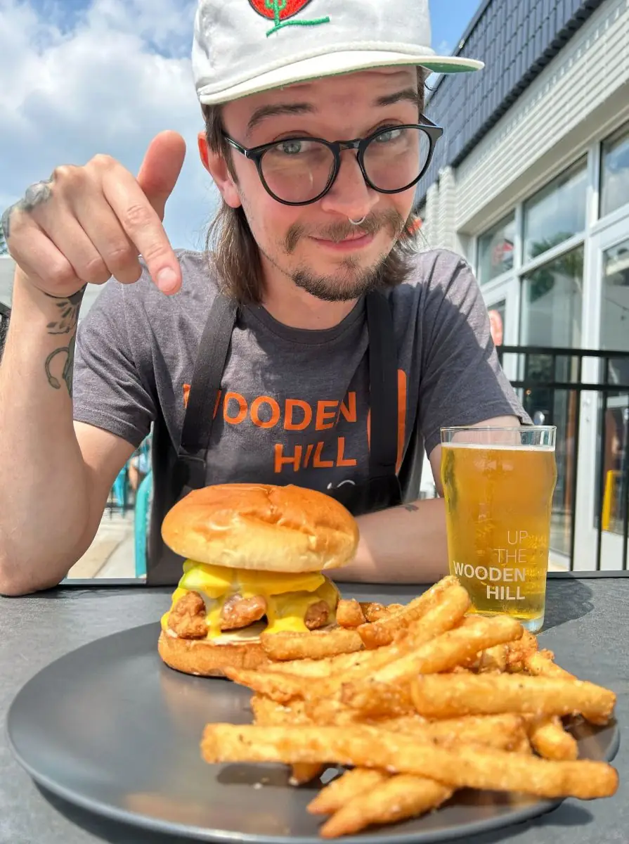 Honey Apple Chicken Sandwich on a plate with fries. Wooden Hill branded glass with a light beer in the background. On a table with blue chairs and the patio in the background.