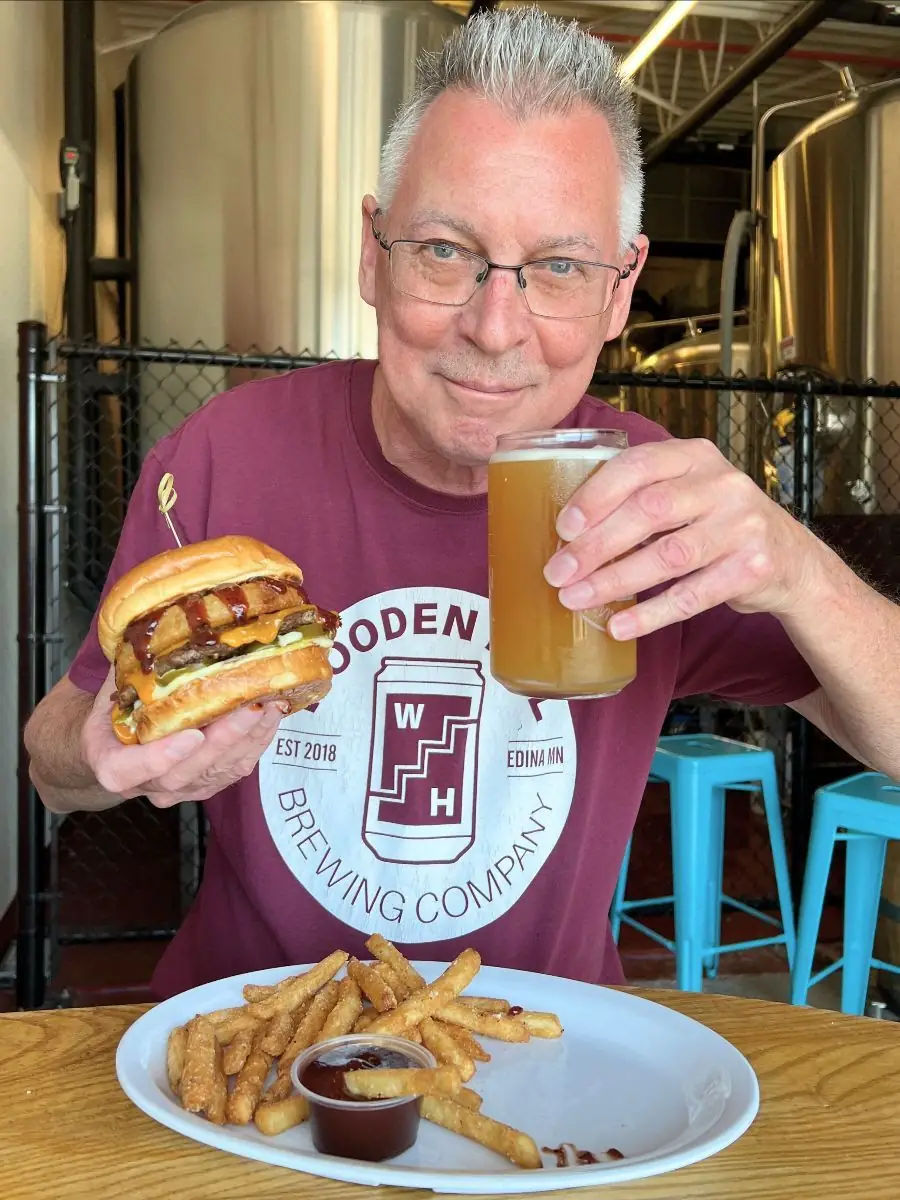 A guy holding the cowboy burger and a beer in his hands. Plate in front of him on the table with fries and BBQ sauce. 