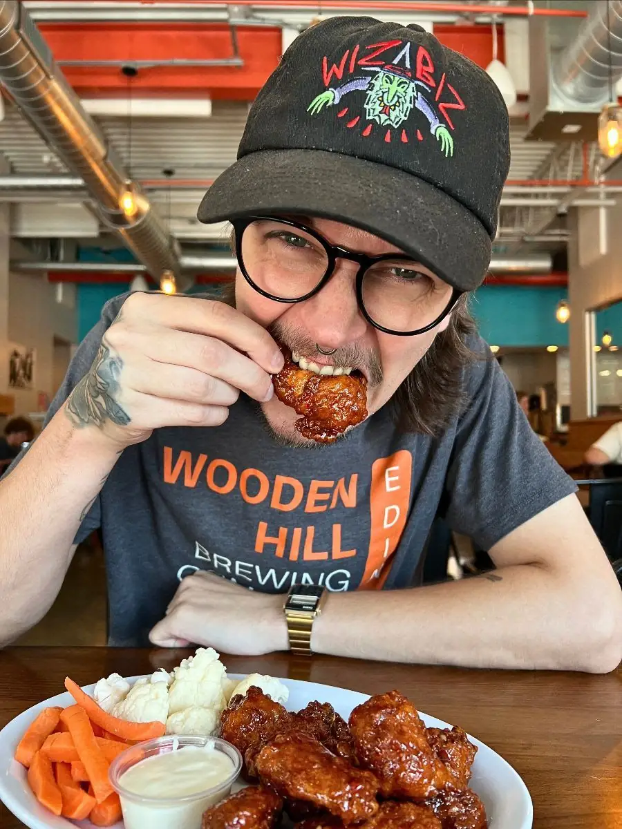 Someone eating a chicken wing with a plate in front of them with wings, cauliflower, carrots, and ranch in a cup.,