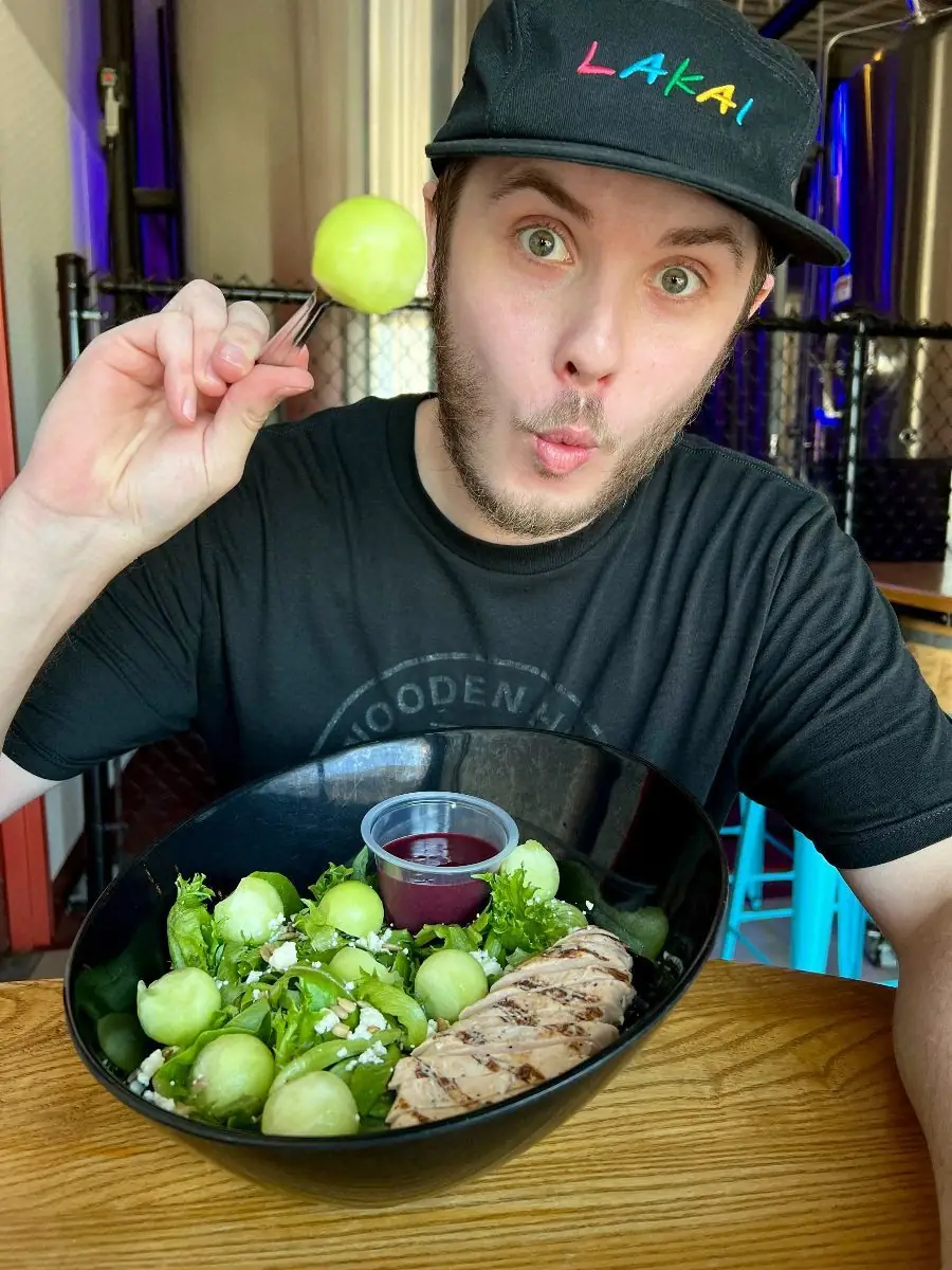Picture of our new Melonberry Crunch Salad! Pictures shows the salad in a black bowl with Kyler, Sous Chef, holding up a melon ball.