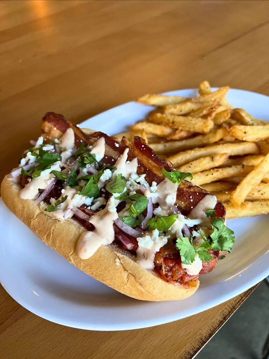 Image of a plate with a ryan dog (hot dog, bacon, queso fresco, roasted salsa, honey chipotle, cilantro, onion) and fries.