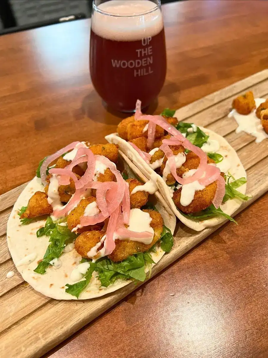 Image of Cheese Curd tacos on a wood board with ranch and cheese curds on the plate in the back. Wooden Hill branded reddish beer in the background on the table.