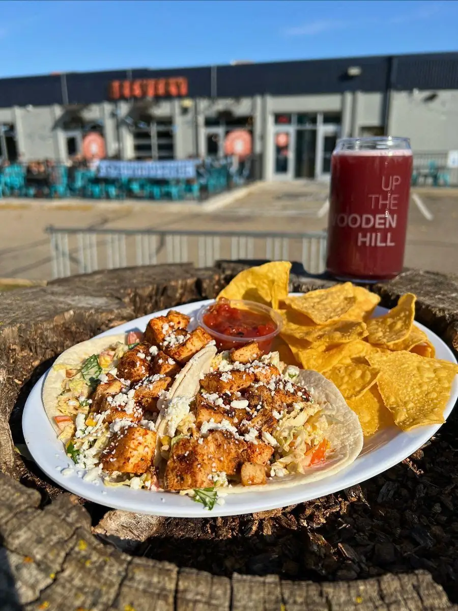 Image of two tequila lime chicken tacos on a plate with chips and salsa. Plate is on a tree stump with a glass of a reddish beer in the background. In the background of the image is the front of Wooden Hill Brewing.
