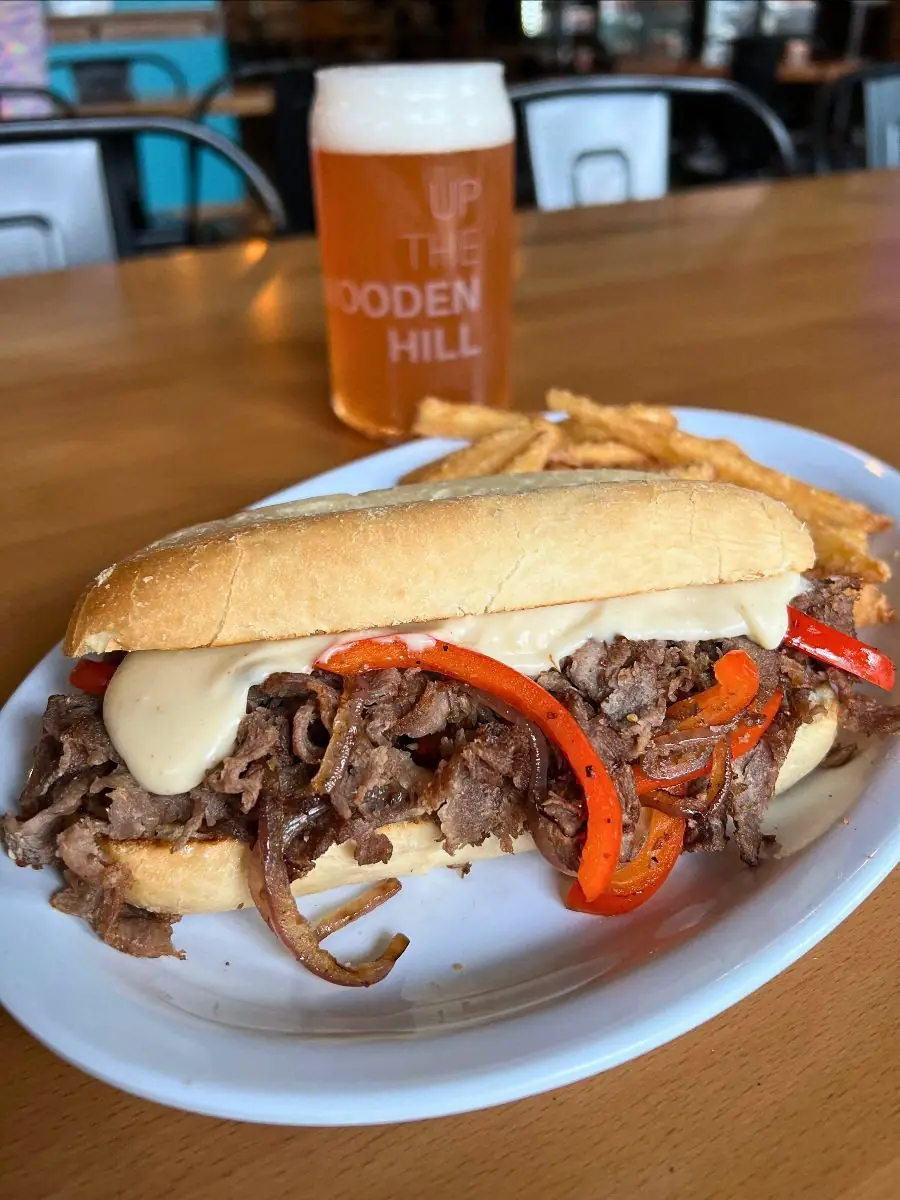 Image of a philly cheesesteak on a plate with french fries. On a wooden table with a Beer in the background.