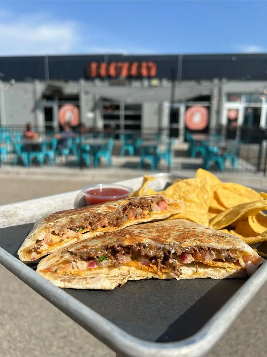 Picture of this week's taco special. This week, we're bringing back a favorite - Crunchy Wrap. Picture shows a crunchy wrap on a sheet pan with a side of chips & salsa. Picture was taken in front of the front patio.