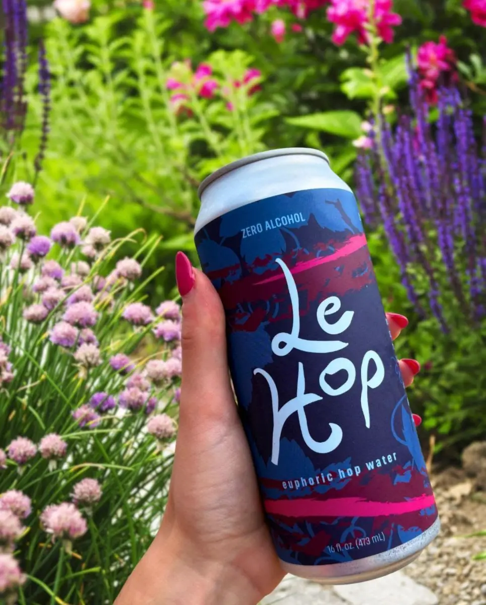 Picture of our new euphoric hop water, Le Hop Moonlight Dark Berry! Picture shows a hand holding a can of Le Hop with pink and purple flowers in the background.