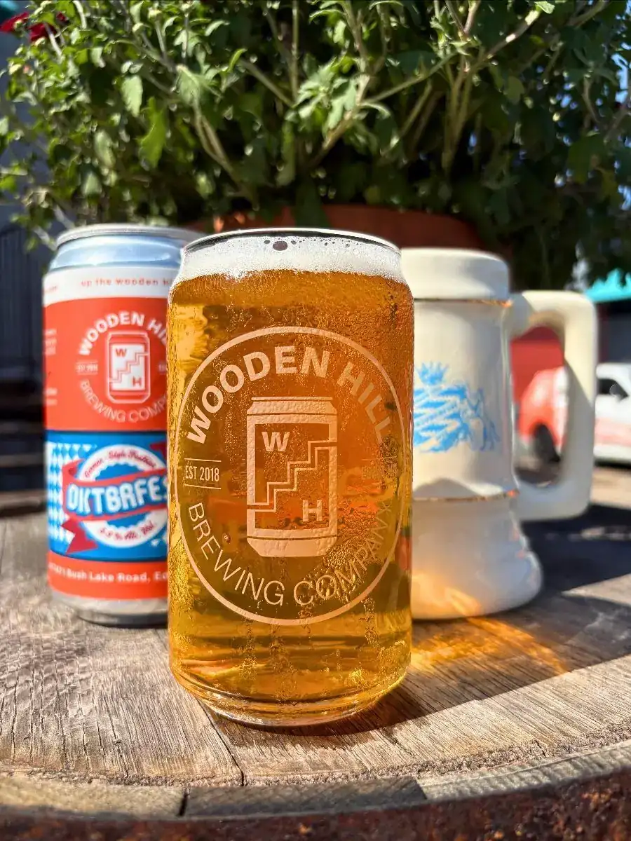 Front of image is a Wooden Hill branded glass with a light beer in it. Behind the glass of beer is a can of Wooden Hill Oktbrfest on the left and a cream colored stein with a blue logo on the right. Plant is in the background.
