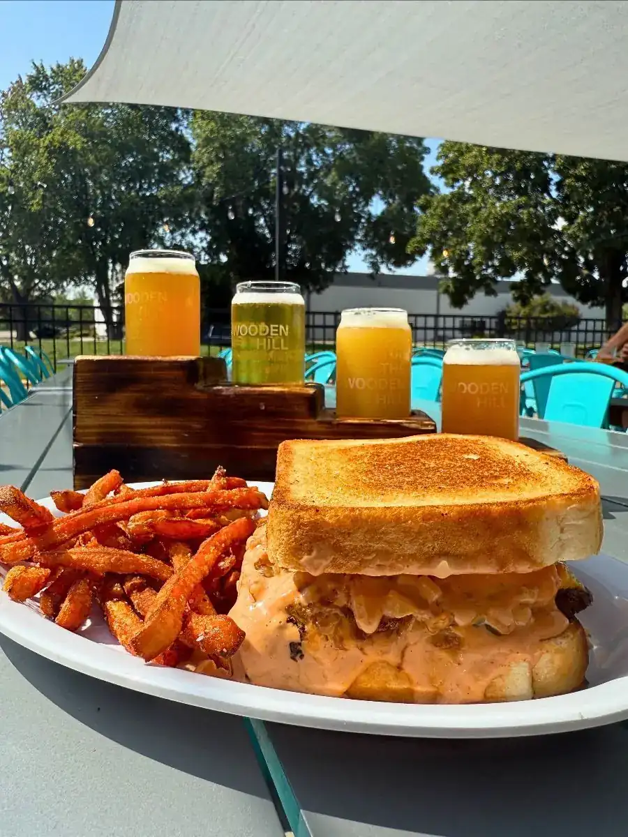 Picture shows the WH Patty melt. Patty melt on a plate with sweet potato fries, flight of beers on a stair shaped flight in the background all on patio with trees in the background.