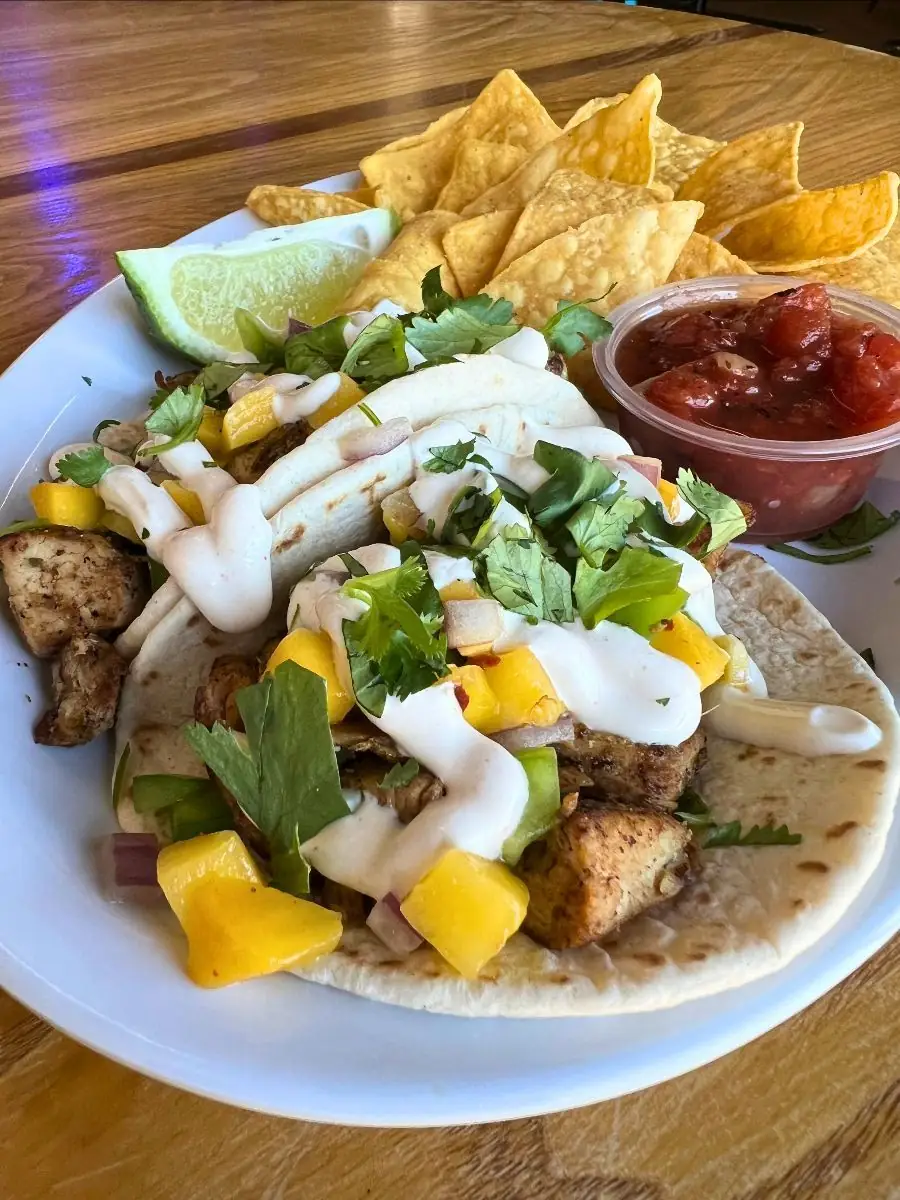 Two soft tacos filled with marinated grilled jerk chicken thighs, sweet mango salsa, mango habanero crema, and cilantro, served with a side of tortilla chips and salsa.