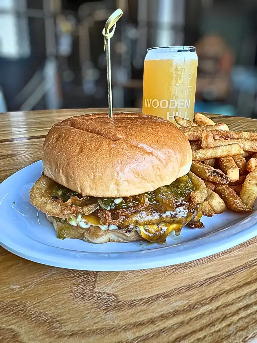 A juicy Angus beef burger topped with cheddar cheese, crispy onion tangles, house garlic mayo, and jalapeño honey jam, served with seasoned fries and a Wooden Hill beer.