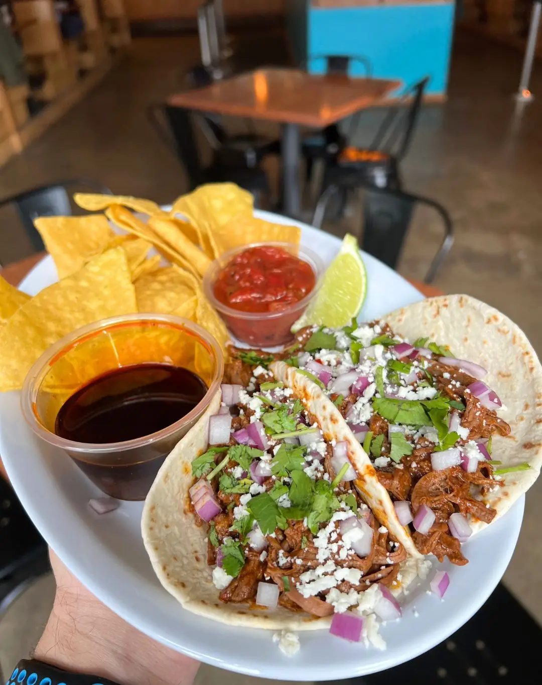 Picture of this week's taco special - Birria Tacos! Picture shows two tacos served with a side of chips & salsa, consomme, and a lime wedge. Tacos are served on a white plate. Picture was taken in the front of our taproom.
