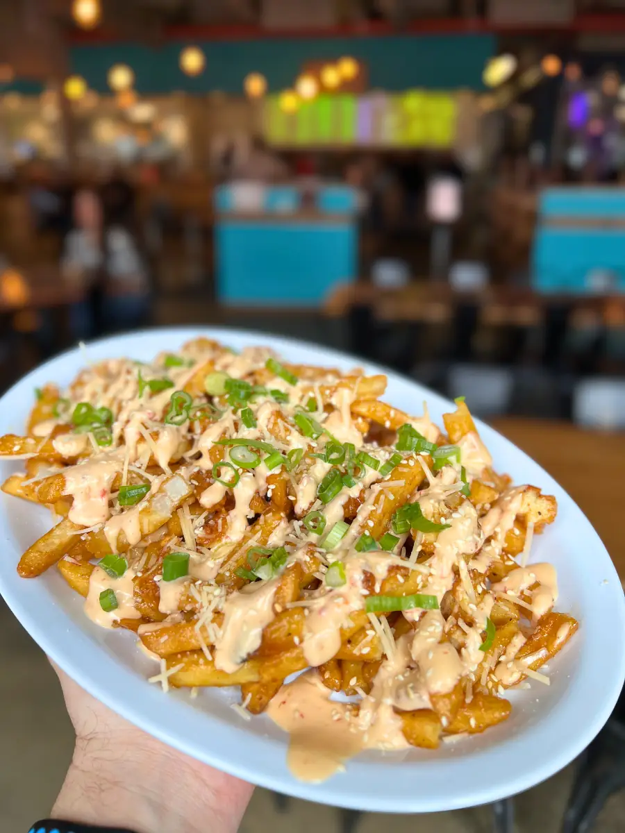 A plate of fries tossed with chopped chicken tenders, and a zesty orange sauce, topped with shredded parmesan cheese, bang bang sauce, green onions, and sesame seeds.