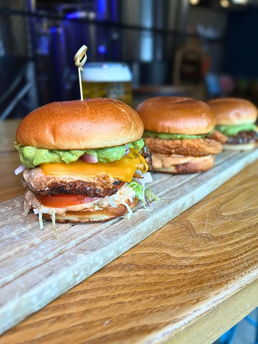 An eye-catching close-up of a stacked California Chicken Sandwich, skewered on a wooden table. Each soft brioche bun is filled with your choice of Grilled or Crispy chicken, melted cheddar, vibrant avocado spread, and smoky chipotle mayo.