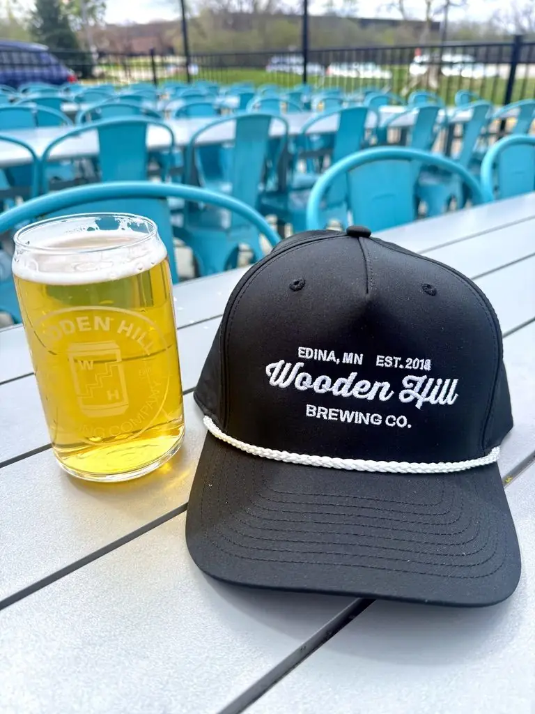Black Hat with Wooden Hill stitched in a cursive white font, white rope along brim of hat. Beer in background.