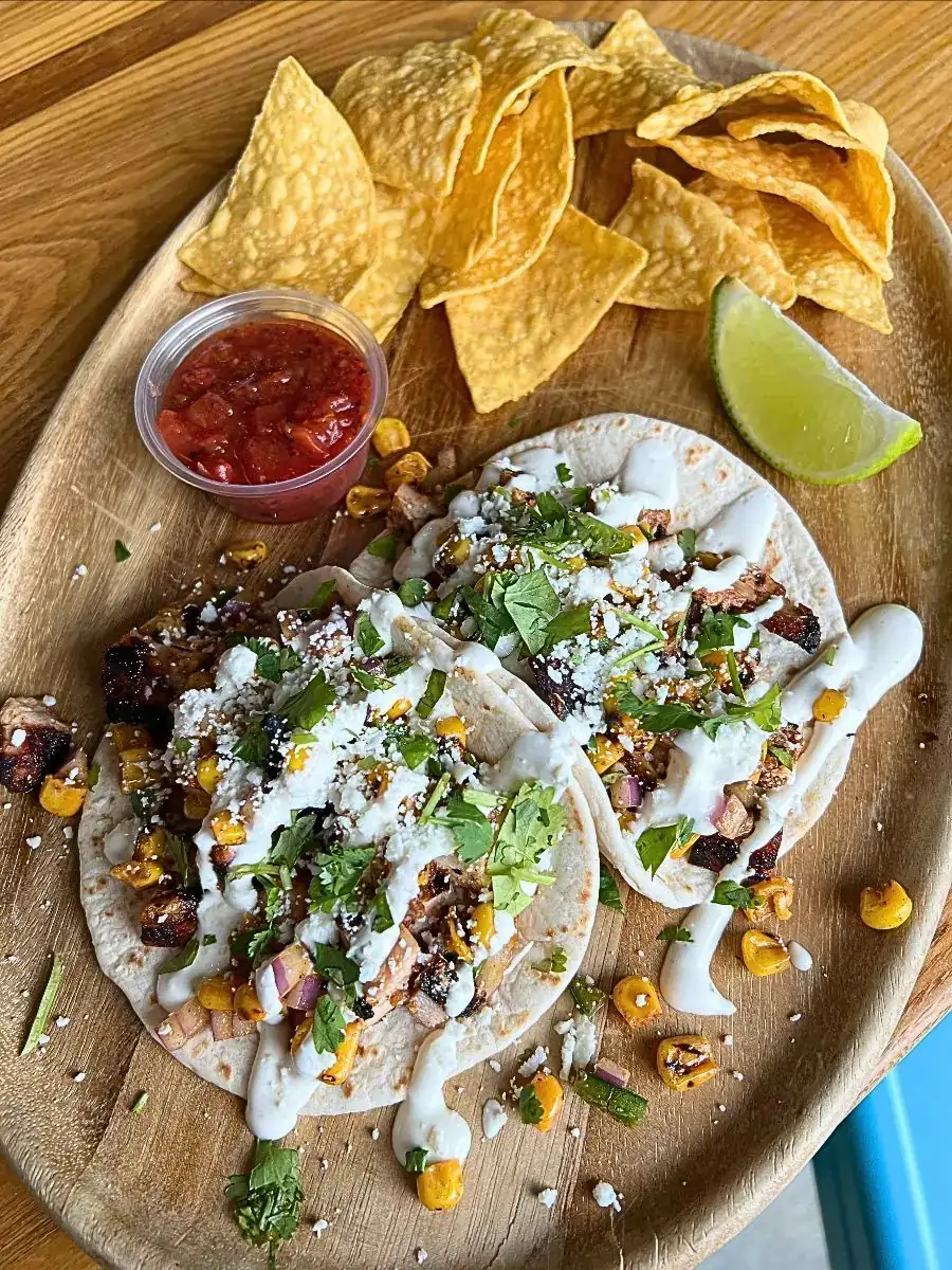 Two adobo grilled chicken tacos on soft flour tortillas, topped with roasted corn salsa, creamy elote sauce, queso fresco, and cilantro. Served on a wooden plate with a lime wedge, tortilla chips, and red salsa.