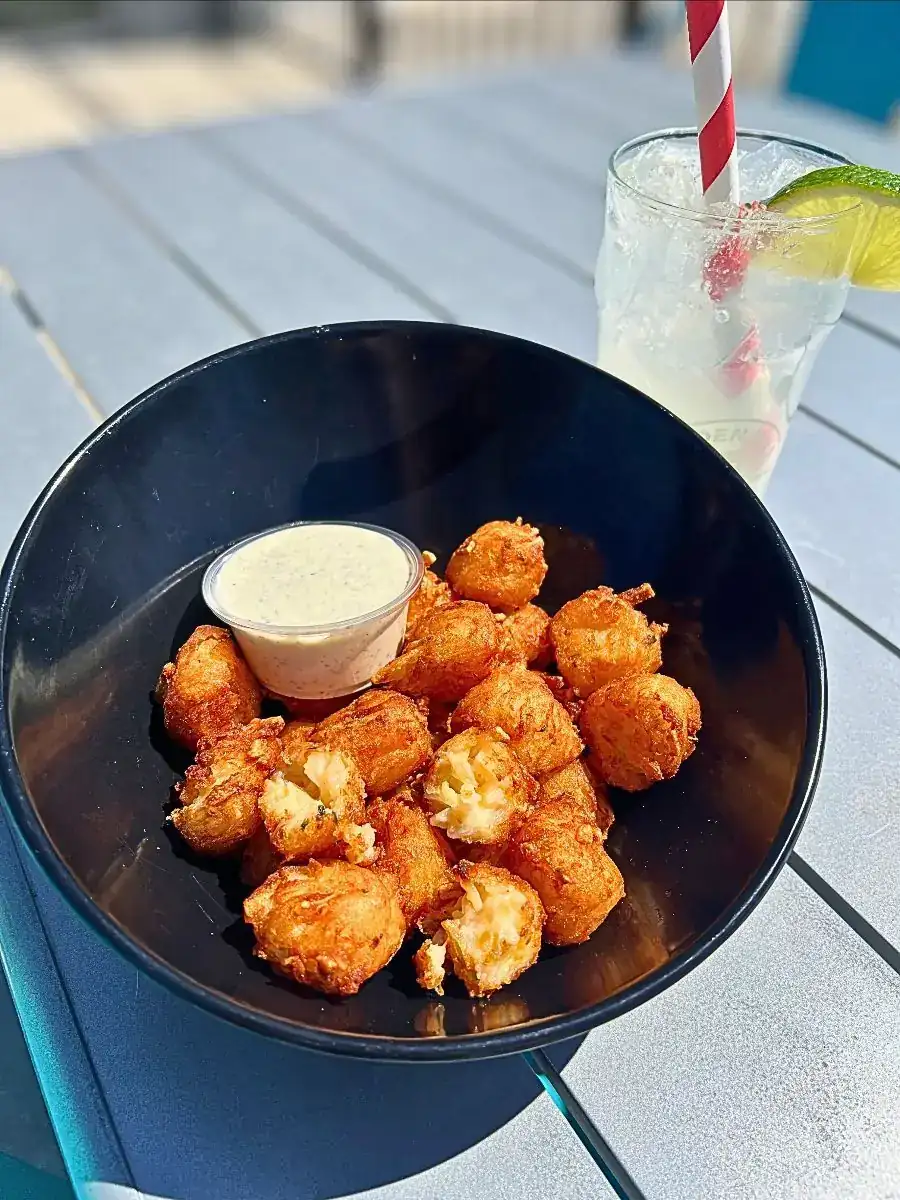A bowl of crispy tater tot nuggets stuffed with sour cream, cheddar, and chive, served with a side of house-made roasted jalapeño ranch. Set on an outdoor table with a refreshing iced drink in the background.