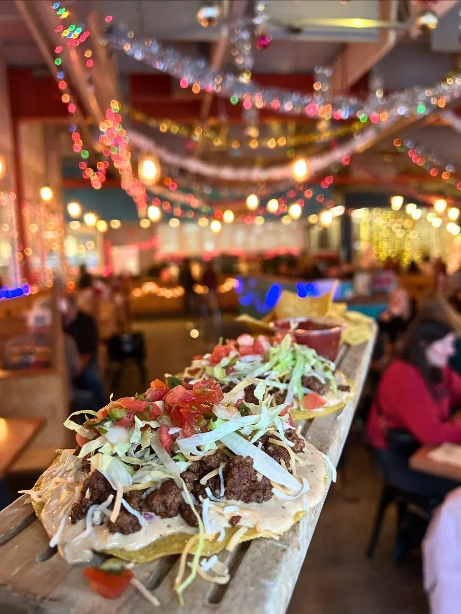 Image of two smash burger tostadas on a wood paddle with chips and salsa. Background is the brewery decorated with tinsel and lights.