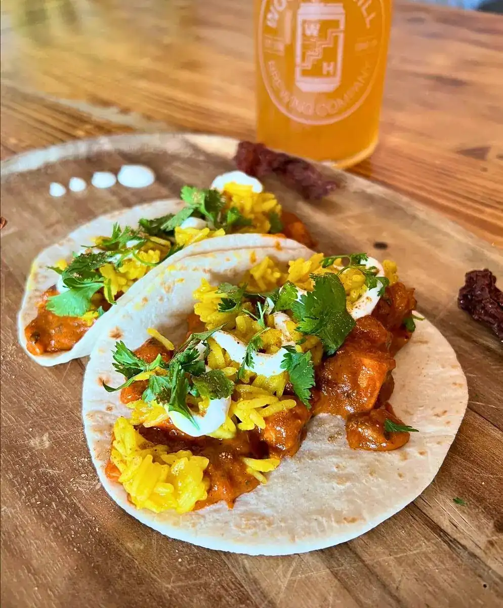 Image of two butter chicken tacos on a wood board with a Wooden Hill glass in the background with a light beer in it.