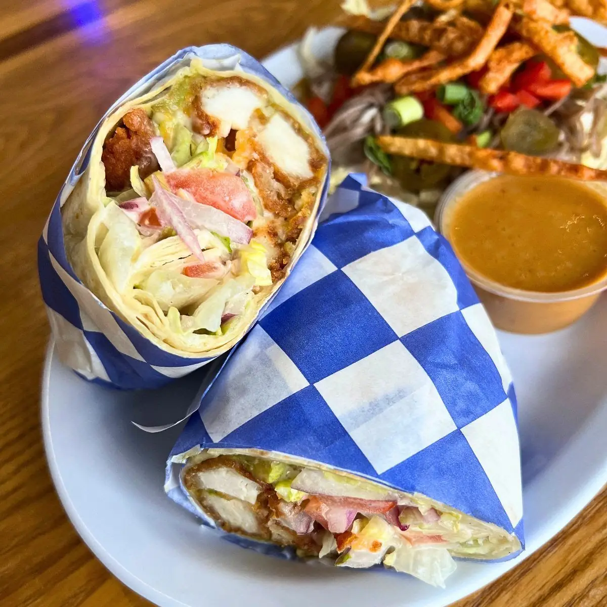 Image of a California wrap wrapped in a blue and white checked wrapping cut in half. Background is of a soba salad on the plate and a brown colored dressing in a cup.