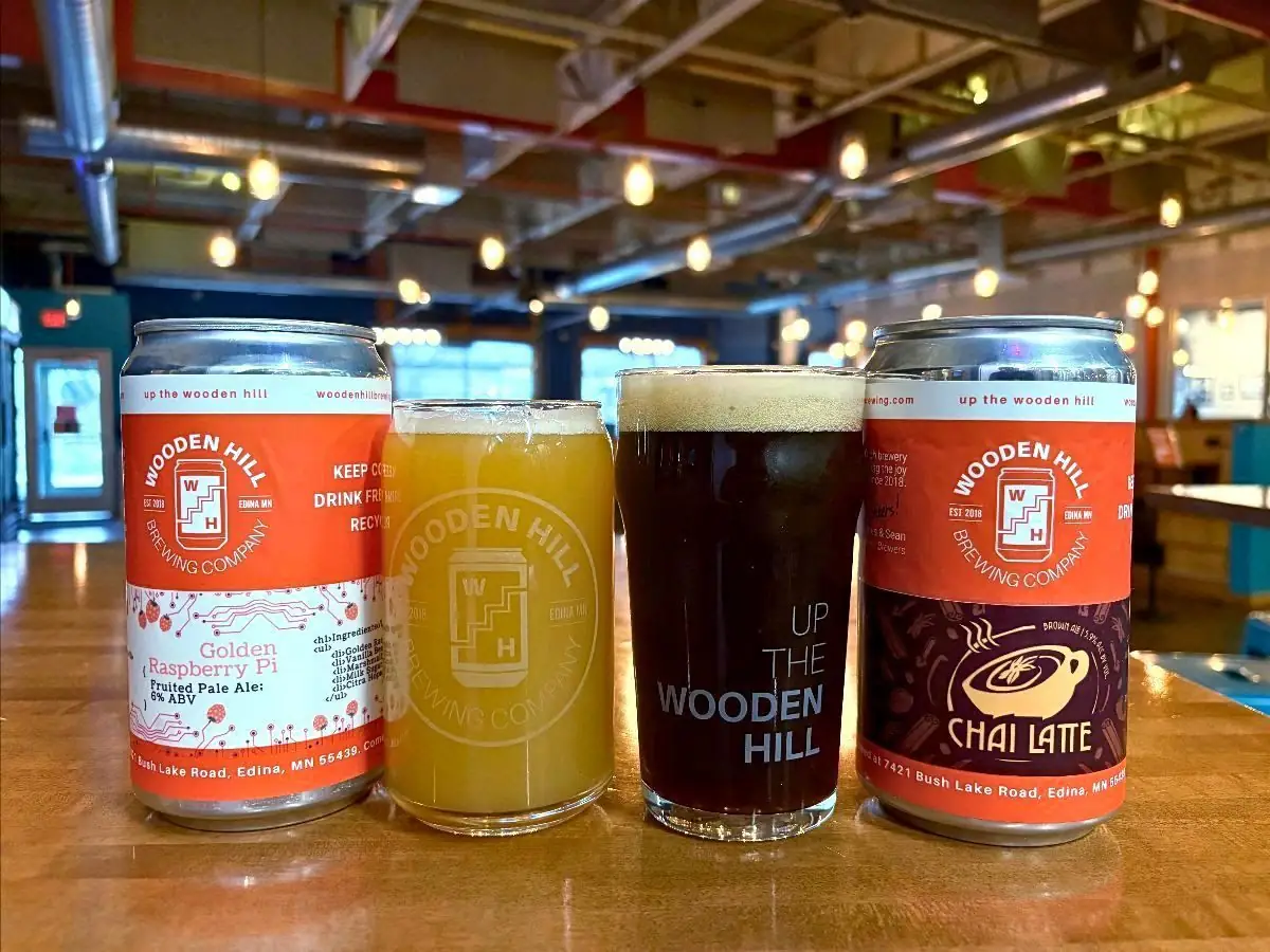 Image of a can of Wooden Hill Golden Raspberry Pie beer on the left with a glass of a hazy beer to the right of it. To the right of that is a glass of a darker malty beer and to the left of that is a Wooden Hill can of Chai Latte. Brewery in the background.
