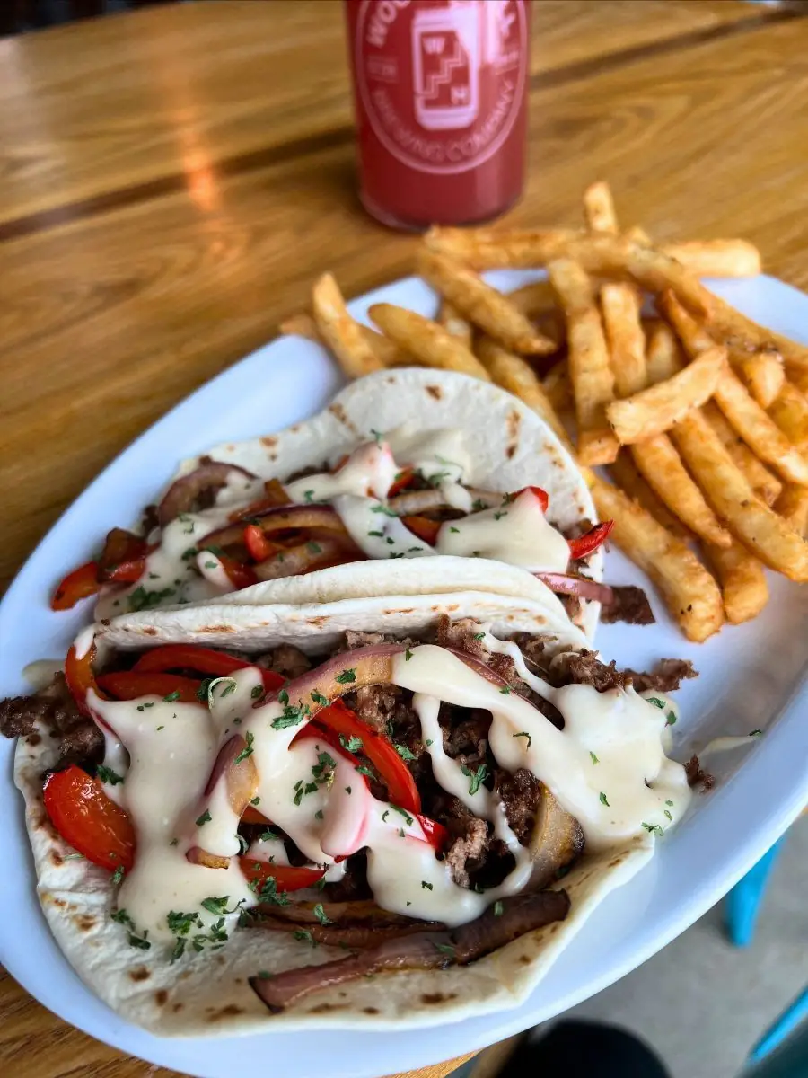 Image of two cheesesteak tacos on a plate with fries. Glass of a rose colored beer in the background all on a wood table.