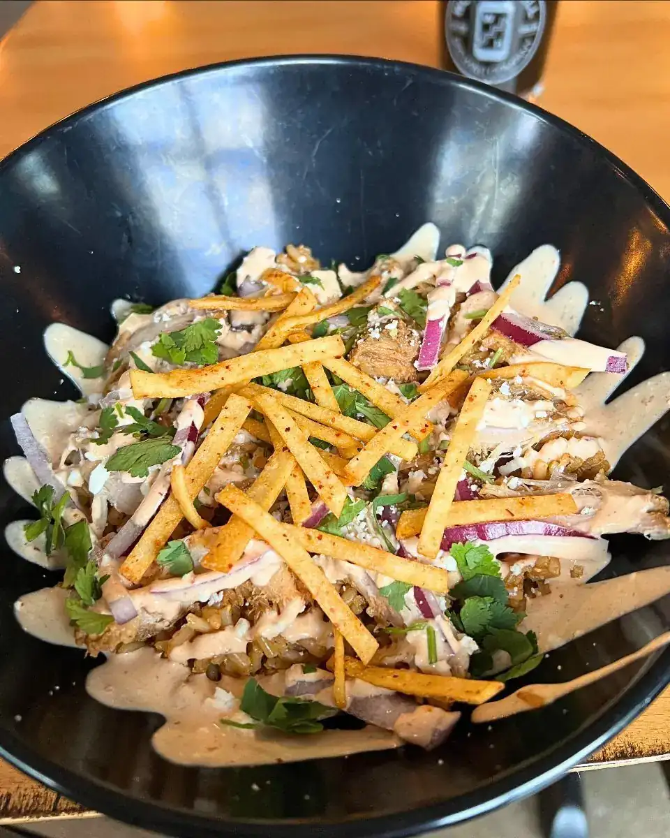 Image of a Carnitas Bowl in a black plastic bowl. There is a glass with a dark beer in the background.