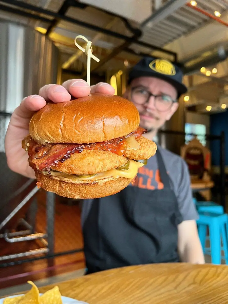 Image is focused on a facemelter fried chicken sandwhich with a hand holding it. Guy in the background holding it in the brewery with a tank in the back.